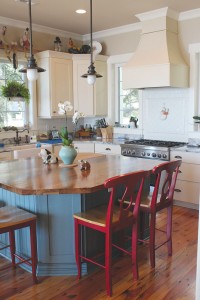 The kitchen remains coastal with its light, airy feel, while accents such as subway tiles, a farmhouse sink, and red bar chairs offer country-chic style. Photo by Bethany Turner