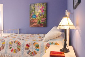 The main house's master bedroom is painted a sweet lavender, matching the quaint quilt and pretty flower painting. Photo by Bethany Turner